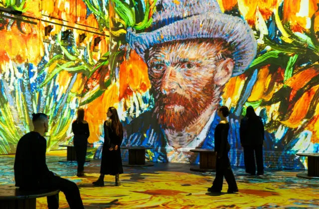 People observing a digital Van Gogh art installation with vibrant colors and large floral projections on the walls and floor.