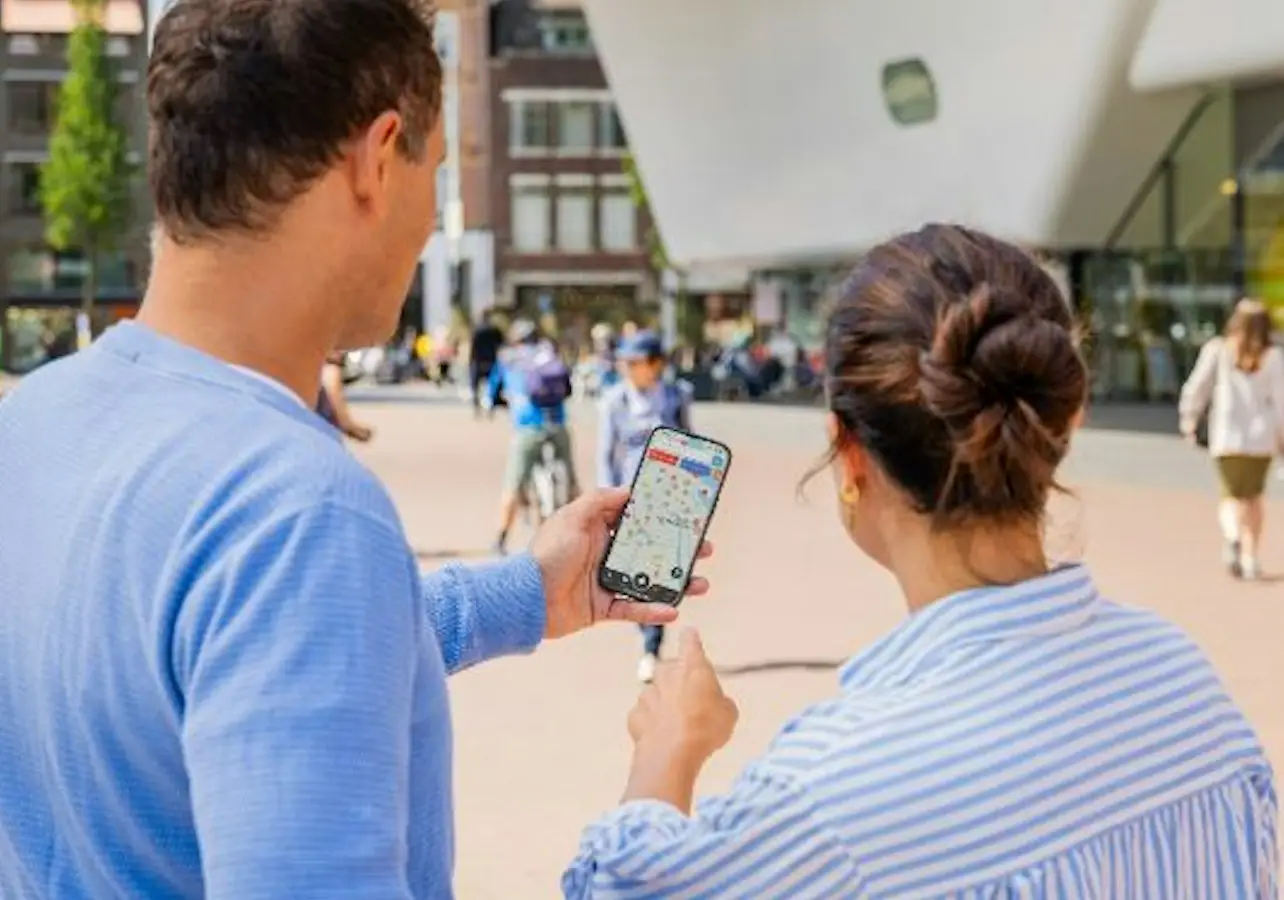 Two people outdoors looking at a smartphone map, with buildings and a modern structure in the background.