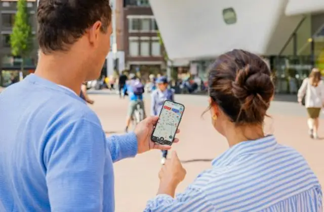 Two people outdoors looking at a smartphone map, with buildings and a modern structure in the background.