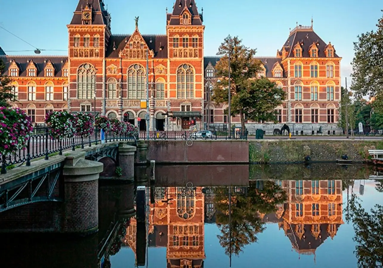 Historic building with ornate architecture reflected in a calm canal, bordered by a bridge and trees under a clear blue sky.
