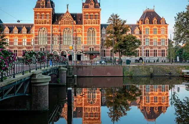 Historic building with ornate architecture reflected in a calm canal, bordered by a bridge and trees under a clear blue sky.