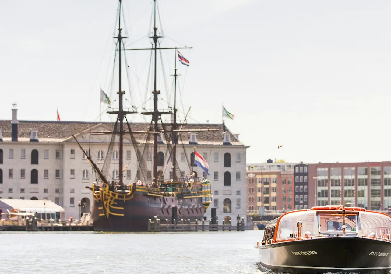 A tourist boat on a canal passes near a docked historic ship and a large building with flags in the background.