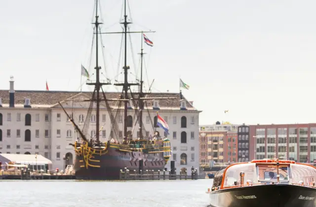 A tourist boat on a canal passes near a docked historic ship and a large building with flags in the background.