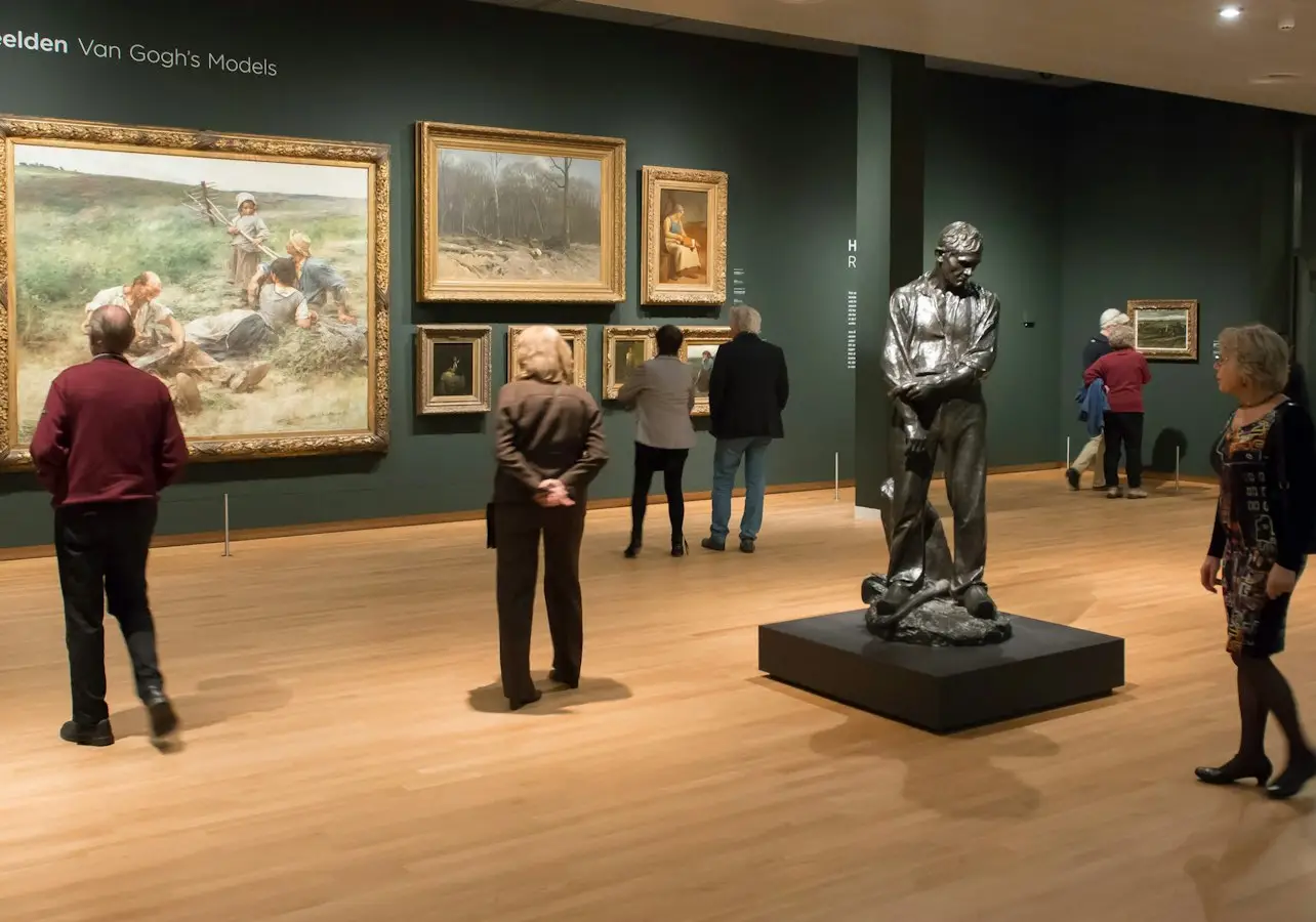 Visitors in an art gallery observing paintings on walls and a statue on a pedestal in a well-lit room.