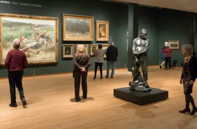 Visitors in an art gallery observing paintings on walls and a statue on a pedestal in a well-lit room.