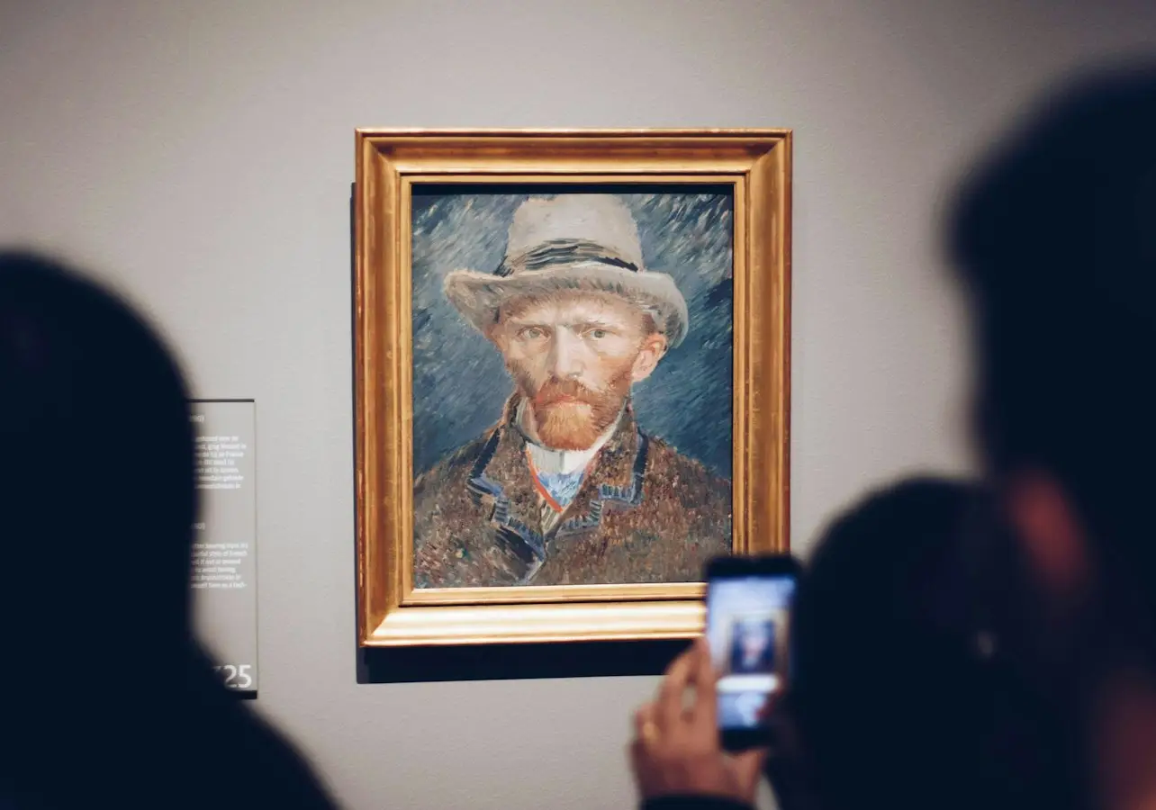Two people viewing and photographing a framed portrait of a man wearing a hat, displayed on a museum wall.