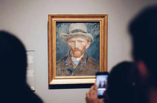 Two people viewing and photographing a framed portrait of a man wearing a hat, displayed on a museum wall.