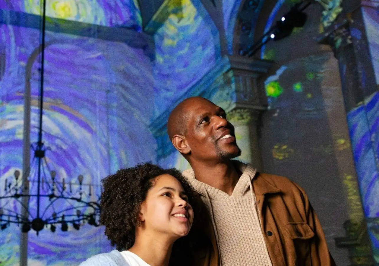 Two individuals standing under colorful projections in an indoor setting with decorative chandeliers and columns.