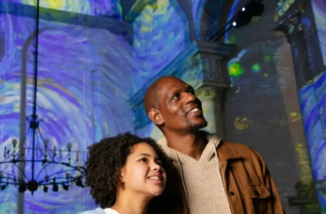 Two individuals standing under colorful projections in an indoor setting with decorative chandeliers and columns.